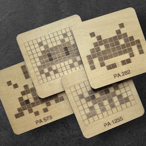 Puede incluir: Cuatro posavasos de madera con diseños de invasores espaciales pixelados. Cada posavasos tiene un diseño único y el texto "PA 573", "PA 1255" y "PA 282" impreso en ellos.