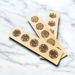 Puede incluir: Tres marcapáginas de madera con diseños grabados con láser. Un marcapáginas presenta un patrón repetido de una flor estilizada, otro presenta un patrón repetido de una hoja estilizada y el tercero presenta un patrón repetido de una flor estilizada.