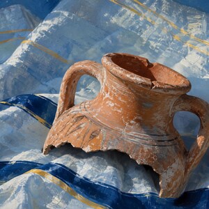 Puede incluir: Una ánfora de terracota rota con dos asas y detalles de líneas pintadas oscuras, sobre una tela a rayas blancas y azules con finos acentos amarillos. El recipiente de cerámica presenta un borde irregular y una base fracturada.