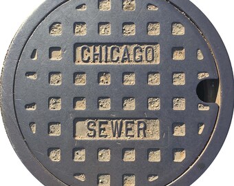 May include: A circular, dark gray manhole cover with the words "CHICAGO" and "SEWER" embossed in rectangular panels. The cover has a grid pattern of square holes and a slightly raised outer rim. The surface shows signs of wear and dirt.