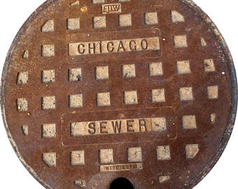 May include: A circular, rusty brown manhole cover with the city name "CHICAGO" and the word "SEWER" in rectangular boxes. The cover has a grid pattern of square holes and a small opening at the bottom. The text "MADE IN USA" is also visible.
