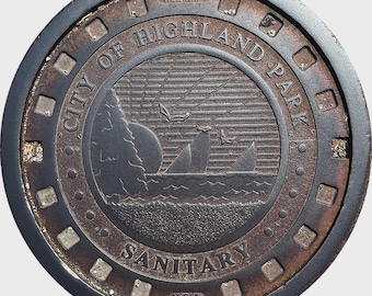 May include: A circular, weathered metal manhole cover. The design features a coastal scene with sailboats, a sun, and the text "CITY OF HIGHLAND PARK SANITARY USA". The cover has a dark outer ring and a detailed inner design.