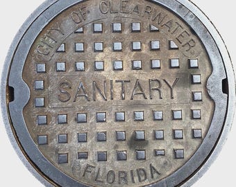 May include: A weathered, circular manhole cover with the text "CITY OF CLEARWATER" and "SANITARY FLORIDA" in a circular pattern. The cover has a grid pattern of small squares and a metallic finish.