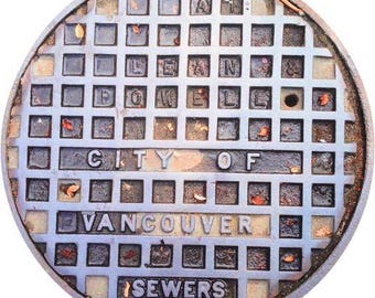 May include: A circular, weathered metal manhole cover with a grid pattern. The words "CITY OF VANCOUVER SEWERS" are prominently displayed. The cover has a dark, aged appearance with some debris.