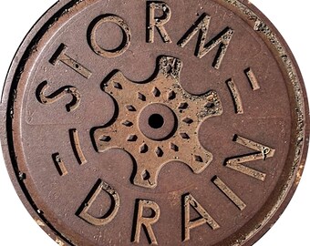 May include: A circular, rusty brown storm drain cover with the words "STORM DRAIN" embossed around a central gear-shaped design. The cover has a weathered appearance, with visible wear and tear.