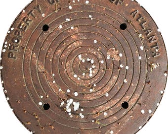 May include: A circular, rusty brown manhole cover with the text "PROPERTY OF CITY OF ATLANTA" and "KOREA" inscribed. The cover features a spiral design and several small holes. White specks and small debris are scattered across the surface.