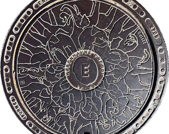May include: A circular, dark metal manhole cover with a detailed relief design of elephants. The cover has a central circular emblem with the letter "E" and text, surrounded by a decorative border with text and rivets. The design is intricate and artistic.