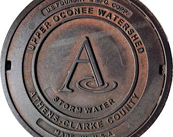 May include: A circular, weathered metal manhole cover. The text "UPPER OCONEE WATERSHED" and "ATHENS-CLARKE COUNTY" are visible. The center features a large "A" above water lines. The cover is marked "MADE IN U.S.A."