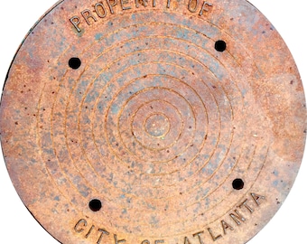 May include: A circular, weathered metal cover with the text "PROPERTY OF" and "CITY OF ATLANTA" in a ring. The cover has concentric circular grooves and four evenly spaced holes. The metal has a rusty brown color with hints of blue.