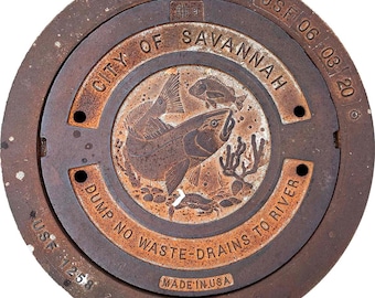 May include: A circular, rusty metal manhole cover with the text "CITY OF SAVANNAH" and "DUMP NO WASTE-DRAINS TO RIVER." The center features an engraved design of fish and aquatic plants. The words "MADE IN USA" are also visible.