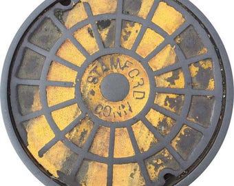 May include: Circular, weathered manhole cover with a grid pattern and the text "STAMFORD CONN". The cover features a central circle with the text and radiating yellow and black segments. The outer rim is dark gray.