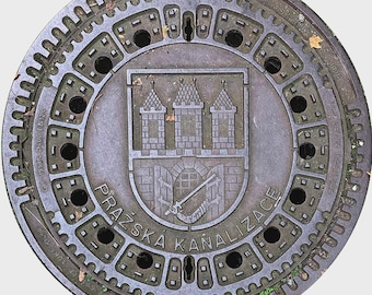 May include: A circular, dark metal manhole cover with a detailed design. The center features a coat of arms with three towers and a sword. The text "PRAZSKA KANALIZACE" is inscribed around the emblem. The cover has a textured, cog-like outer rim.