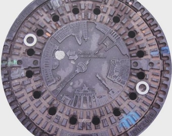 May include: A circular, dark gray manhole cover with a detailed relief design. The design includes the text "BUDERUS" and a depiction of Berlin landmarks, including the Brandenburg Gate and the TV Tower. The words "Berliner Wasserbetriebe" are also visible.