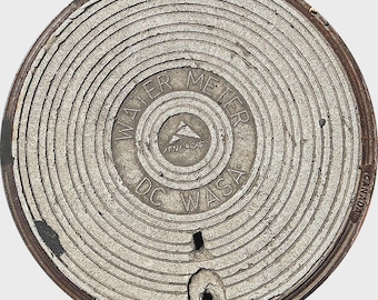 May include: Circular, weathered metal water meter cover with concentric grooves. The text "WATER METER" and "DC WASA" are visible, along with a logo. The cover has a dark, rusty edge and is marked "CANADA".