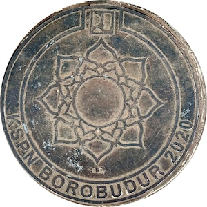 May include: A weathered, circular metal object with a central floral design and the text "KSPN BOROBUDUR 2020" around the edge. The design features a central circle surrounded by a stylized flower pattern. The object has a worn, aged appearance.