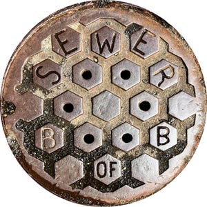 Puede incluir: Una tapa de alcantarilla metálica circular y desgastada con las palabras "SEWER BOARD OF" en relieve. El diseño presenta un patrón de panal con agujeros circulares. La tapa tiene un aspecto oxidado y envejecido.