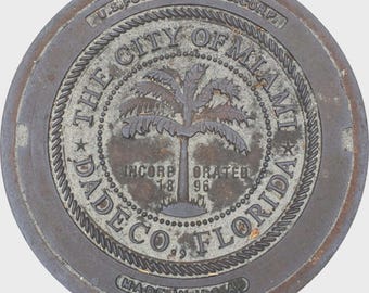 May include: A weathered, circular metal plate with the text "THE CITY OF MIAMI FLORIDA" encircling a palm tree design. The plate also includes the text "INCORP ORATED 18 96" and "MADE IN U.S.A."