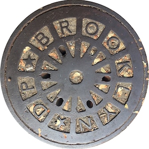 May include: A close-up of a round, black metal manhole cover with the word "BROOKLYN" spelled out in raised letters around the edge. The cover is decorated with a circular pattern of small, triangular shapes.