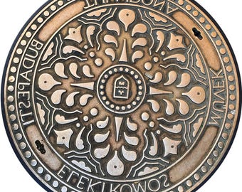 May include: A circular, bronze-colored manhole cover with an ornate, symmetrical design. The cover features a central emblem surrounded by stylized floral and geometric patterns. The outer rim has text in a circular pattern, with the words "BUDAPESTI" and "EFEKTIKOWOZ" visible.