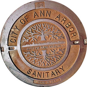 Könnte beinhalten: Ein rostiger Metallschachtdeckel mit dem Text "CITY OF ANN ARBOR" und "SANITARY" in einem Kreis um einen Baum mit Wurzeln und einer Stadtlandschaft. Der Text "EST. CITY OF ANN ARBOR 1824" befindet sich unter dem Baum. Der Text "MADE IN USA" befindet sich am unteren Rand des Deckels.