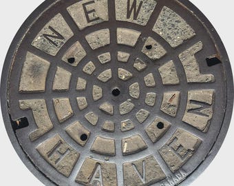 May include: Circular, weathered metal manhole cover with the words "NEW HAVEN" arranged in a radial pattern. The cover has a dark, aged appearance with visible wear and tear, and the text is in a blocky font.