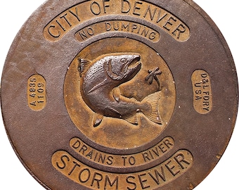 May include: A circular, bronze-colored manhole cover with the text "CITY OF DENVER" and "STORM SEWER". A central emblem features a fish and dragonfly. The cover also includes the text "DRAINS TO RIVER", "NO DUMPING", and "D&L FDRY USA".