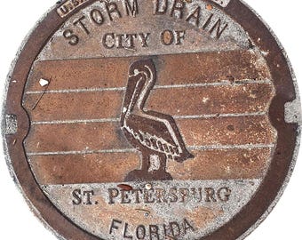 May include: A weathered, circular storm drain cover from St. Petersburg, Florida, with a pelican emblem. The cover is made in the USA and features the text "STORM DRAIN" and "CITY OF". The design includes horizontal lines and a textured surface.
