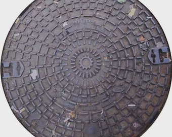 May include: A circular, weathered, gray metal manhole cover with a repeating geometric pattern. The cover has a central circular design and a series of concentric rings. Two blue metal handles are visible on the sides. Some debris is scattered across the surface.