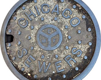 May include: A circular, weathered metal manhole cover with the words "CHICAGO SEWERS" in a bold, uppercase font. The center features a circular emblem with the letters "C", "M", and "F". The cover has a textured surface and is surrounded by a dark metal rim.