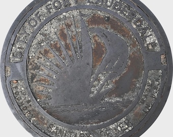 May include: A circular, weathered metal manhole cover. The design features a stylized sunburst over water, with the text "CITY OF FORT LAUDERDALE SANITARY SEWER". The cover has a textured, speckled background and a dark, aged appearance.