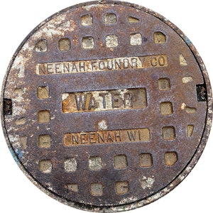May include: A rusty metal water utility cover with the text "NEENAH FOUNDRY CO" and "WATER" in white lettering. The cover also has the text "NEENAH WI" in white lettering.