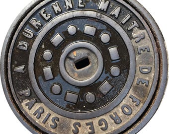 May include: Circular, cast-iron manhole cover with the text "A DURENNE MAITRE DE FORGES PARIS" in a raised, serif font. The cover has a central keyhole and a series of geometric shapes and circles. The color is dark gray with some areas appearing wet.