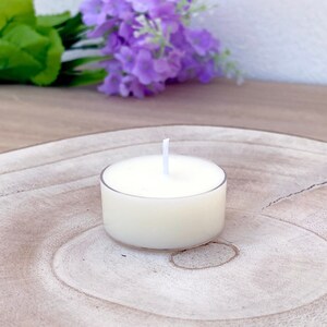 Puede incluir: Una vela de té blanca en un recipiente de vidrio transparente sobre una superficie de madera. La vela está rodeada de un posavasos de madera marrón claro.
