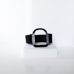 May include: A black, rectangular, adjustable strap with a square frame. The frame is connected to the strap and appears to be made of a hard plastic material. The strap is made of a soft, flexible material.