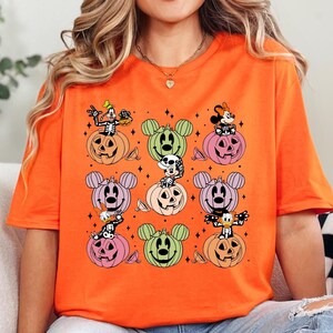 May include: Orange t-shirt with a graphic print of Mickey Mouse, Minnie Mouse, Goofy, and Pluto dressed in Halloween costumes sitting on pumpkins.