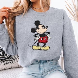 Puede incluir: Una sudadera gris con una imagen de dibujos animados de Mickey Mouse con un atuendo rojo y amarillo. La sudadera tiene mangas largas y cuello redondo.