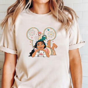 Może przedstawiać: Biały T-shirt z kresk&oacute;wkową postacią Moany z filmu Disneya Moana. Moana leży z swoim domowym prosiaczkiem Puą i kogutkiem Hei Hei. Obrazek jest otoczony sylwetką ucha Myszki Miki z fajerwerkami i sercami.