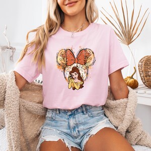 Pu&ograve; includere: Una maglietta rosa con un'illustrazione ad acquerello di Belle de La Bella e la Bestia con una fascia per capelli a forma di orecchie di Topolino. La fascia &egrave; rossa con pois bianchi e un grande fiocco rosso.