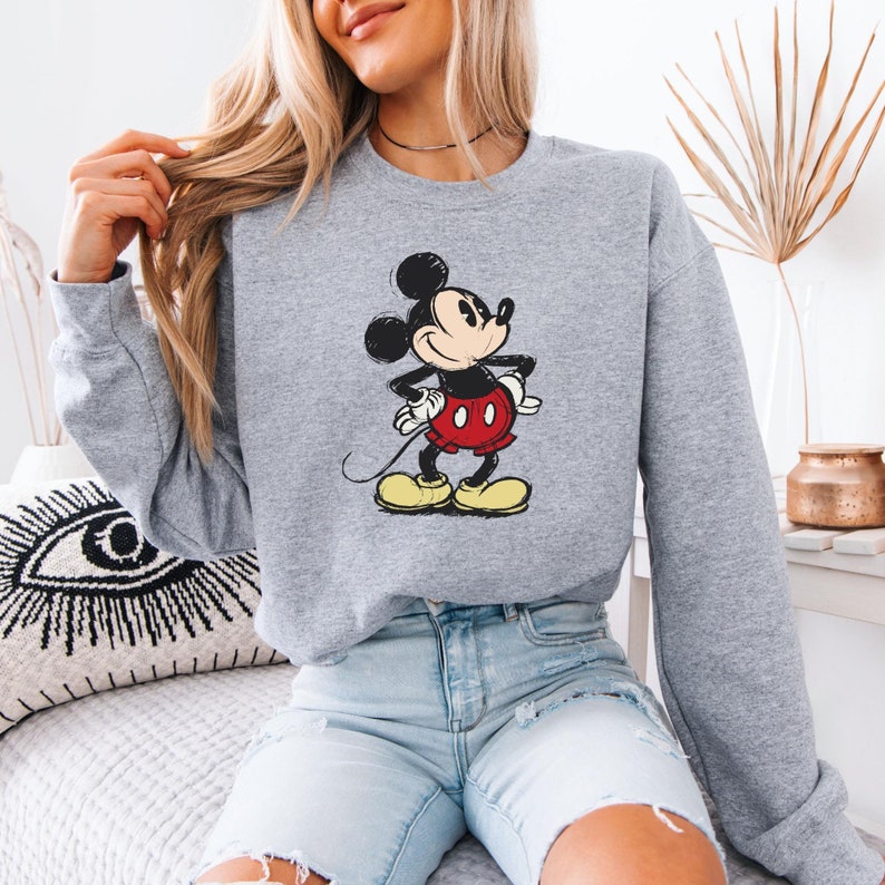 Puede incluir: Una sudadera gris con una imagen de dibujos animados de Mickey Mouse con un atuendo rojo y amarillo. La sudadera tiene mangas largas y cuello redondo.