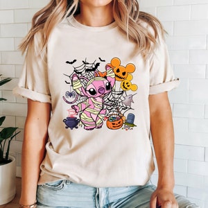 Puede incluir: Una camiseta beige claro con un personaje de dibujos animados rosa vestido de momia. El personaje est&aacute; rodeado de im&aacute;genes de tem&aacute;tica de Halloween, incluyendo murci&eacute;lagos, telara&ntilde;as, un fantasma, un caldero, una calabaza y caramelos.
