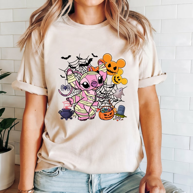 Puede incluir: Una camiseta beige claro con un personaje de dibujos animados rosa vestido de momia. El personaje est&aacute; rodeado de im&aacute;genes de tem&aacute;tica de Halloween, incluyendo murci&eacute;lagos, telara&ntilde;as, un fantasma, un caldero, una calabaza y caramelos.