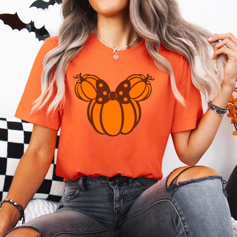 K&ouml;nnte beinhalten: Orangefarbenes T-Shirt mit einem K&uuml;rbis-Schleifen-Design in Orange- und Braunt&ouml;nen. Das kurz&auml;rmelige Shirt ist aus weichem Material. Ein lustiges Halloween-Thema.