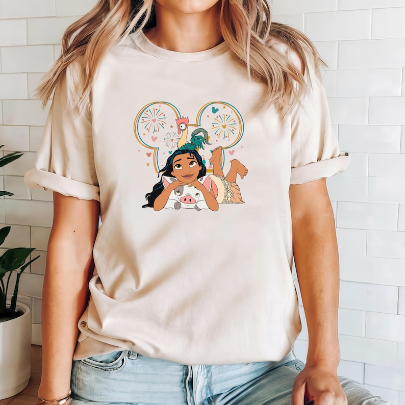 Może przedstawiać: Biały T-shirt z kresk&oacute;wkową postacią Moany z filmu Disneya Moana. Moana leży z swoim domowym prosiaczkiem Puą i kogutkiem Hei Hei. Obrazek jest otoczony sylwetką ucha Myszki Miki z fajerwerkami i sercami.