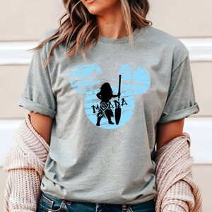 May include: A light gray t-shirt with a blue and white distressed Mickey Mouse silhouette featuring a black silhouette of Moana holding a paddle.