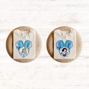 Pu&ograve; includere: Due magliette bianche con un design ad acquerello blu e bianco di una principessa Disney con un fiocco blu. La principessa a sinistra &egrave; Cenerentola e la principessa a destra &egrave; Biancaneve.