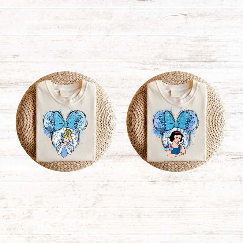 Pu&ograve; includere: Due magliette bianche con un design ad acquerello blu e bianco di una principessa Disney con un fiocco blu. La principessa a sinistra &egrave; Cenerentola e la principessa a destra &egrave; Biancaneve.