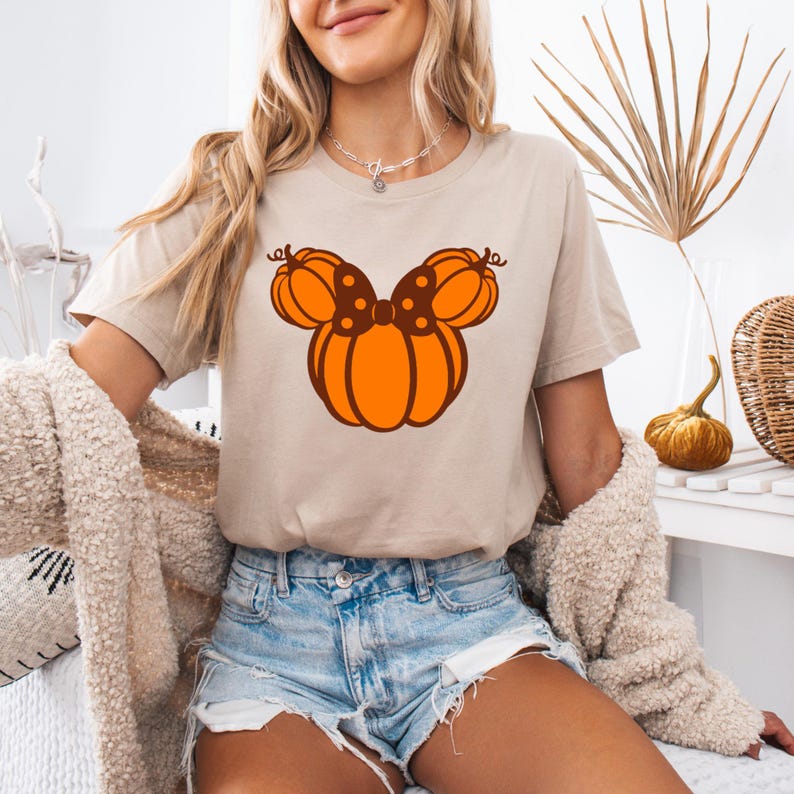 K&ouml;nnte beinhalten: Beiges T-Shirt mit einem orangefarbenen K&uuml;rbis-Design in Form eines Kopfes einer Figur und einer Schleife. Das Design ist mittig auf dem Shirt platziert. Das Shirt wird mit Jeansshorts getragen.