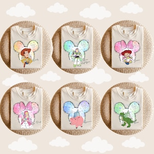 Puede incluir: Seis camisetas blancas con diferentes personajes de Disney en una silueta de oreja de Mickey. Los personajes son Woody, Buzz Lightyear, Jessie, Bella, Hamm y Rex. Cada camiseta tiene el nombre del personaje escrito debajo de la silueta.