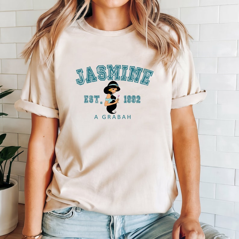 May include: A beige t-shirt with a blue graphic print of Jasmine from Aladdin. The graphic includes the text "Jasmine Est. 1992 A Grabah".