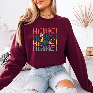 Puede incluir: Una sudadera color burdeos con un gr&aacute;fico de un gallo y el texto "HEIHEI" repetido cuatro veces en estilo retro.